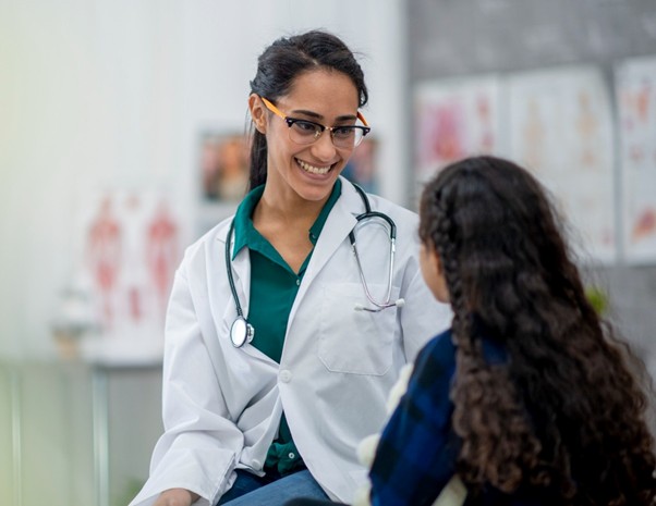A smiling nurse practitioner wearing a white coat talks with a young patient in an examination room.
