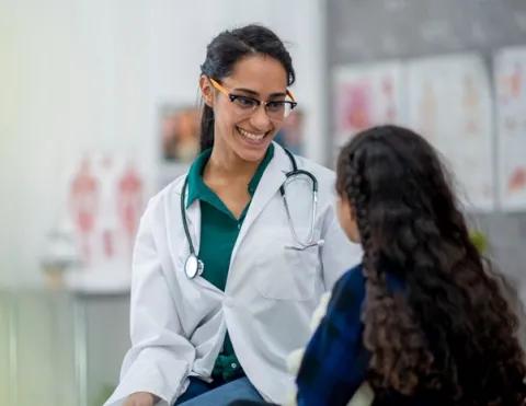 A smiling nurse practitioner wearing a white coat talks with a young patient in an examination room.